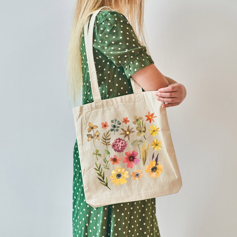 May include: A beige tote bag with a floral design featuring a variety of colorful flowers and leaves. The bag has two long straps and is being held by a person wearing a green polka dot dress.