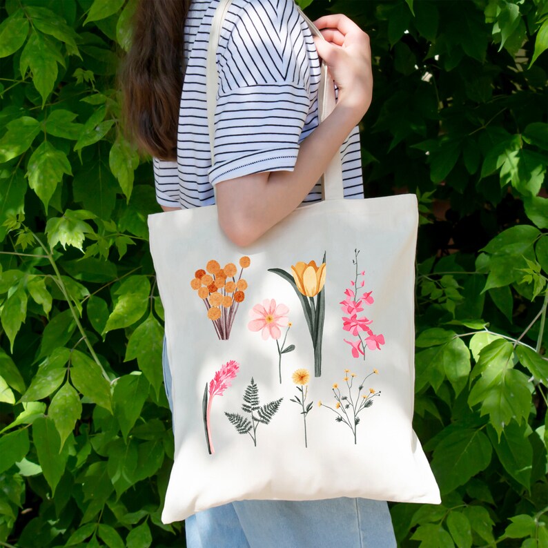 May include: A white canvas tote bag with a floral design. The flowers are pink, yellow, and orange. The bag has two straps.