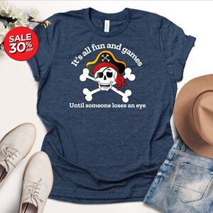 May include: A navy blue t-shirt with a white skull and crossbones graphic. The skull is wearing a red bandana and a pirate hat. The text "It's all fun and games Until someone loses an eye" is printed below the graphic.