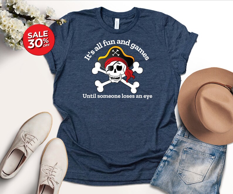 May include: A navy blue t-shirt with a white skull and crossbones graphic. The skull is wearing a red bandana and a pirate hat. The text "It's all fun and games Until someone loses an eye" is printed below the graphic.