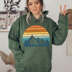 May include: A green hooded sweatshirt featuring a vintage-style graphic of a mountain range and sunset. The graphic has a faded, distressed look with orange, yellow, blue, and green colours.