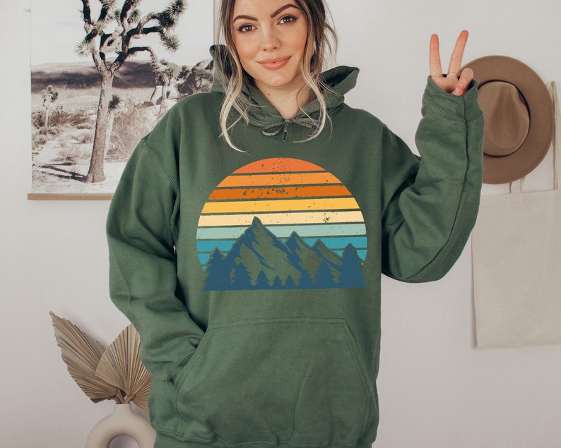 May include: A green hooded sweatshirt featuring a vintage-style graphic of a mountain range and sunset. The graphic has a faded, distressed look with orange, yellow, blue, and green colours.