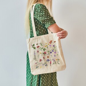 Peut inclure: Une personne tient un sac fourre-tout beige avec un imprim&eacute; floral. Le sac pr&eacute;sente une vari&eacute;t&eacute; de fleurs et de feuilles color&eacute;es imprim&eacute;es dessus. Le sac est en toile et poss&egrave;de deux longues anses.