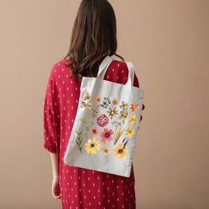 May include: A white tote bag with a floral design featuring various flowers and leaves in shades of yellow, orange, red, and brown. The bag has a long strap and is made of a natural fabric.