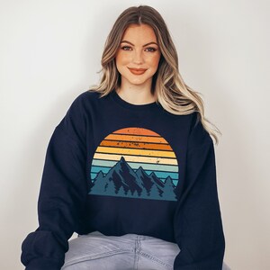 May include: A navy blue sweatshirt featuring a vintage-style graphic of mountains and a sunset. The graphic is in a range of colours, including orange, yellow, blue, and green.