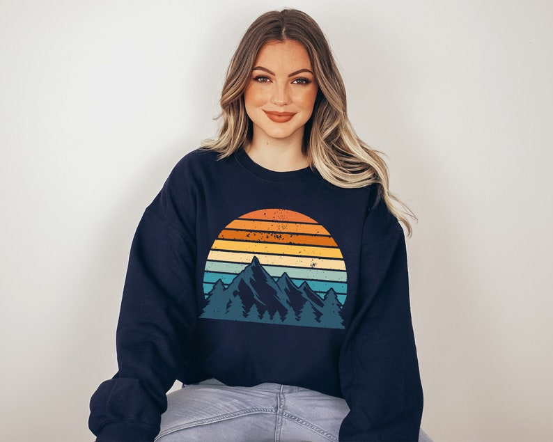 May include: A navy blue sweatshirt featuring a vintage-style graphic of mountains and a sunset. The graphic is in a range of colours, including orange, yellow, blue, and green.
