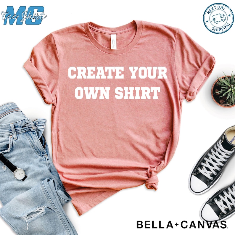 Puede incluir: Camiseta de color rosa polvoriento con el texto blanco "CREATE YOUR OWN SHIRT". La imagen incluye vaqueros azules, un reloj y zapatillas de lona negras. La camiseta es de Bella + Canvas.