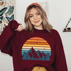 May include: A maroon sweatshirt featuring a vintage-style graphic of a mountain range and a sunset. The graphic is in shades of blue, orange, yellow, and red.