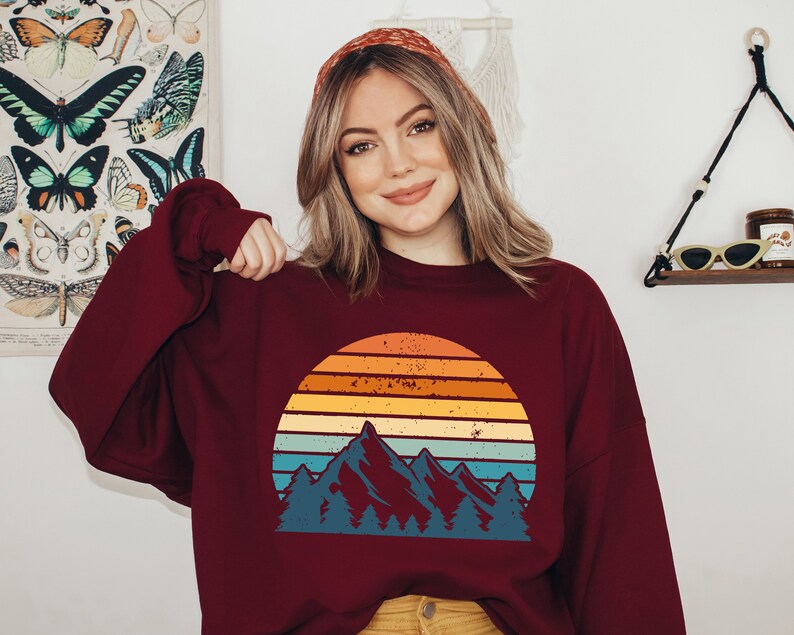 May include: A maroon sweatshirt featuring a vintage-style graphic of a mountain range and a sunset. The graphic is in shades of blue, orange, yellow, and red.