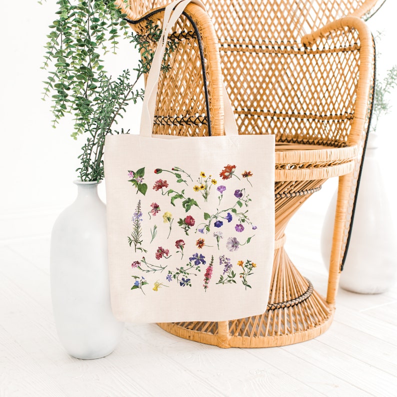 Peut inclure: Un sac fourre-tout en toile blanche avec un imprim&eacute; floral pr&eacute;sentant une vari&eacute;t&eacute; de fleurs color&eacute;es. Le sac a deux longues poign&eacute;es et est pos&eacute; sur une chaise en osier.