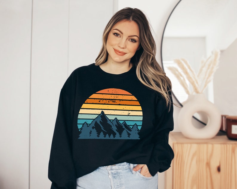 May include: A black sweatshirt featuring a vintage-style graphic of a mountain range and a sunset. The graphic has a faded, distressed look with a rainbow colour palette.