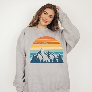 May include: A grey sweatshirt featuring a vintage-style graphic of a mountain range and sunset. The graphic depicts a blue mountain range with trees in front of a yellow, orange, and blue sunset.