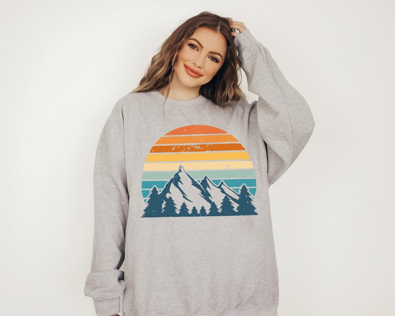 May include: A grey sweatshirt featuring a vintage-style graphic of a mountain range and sunset. The graphic depicts a blue mountain range with trees in front of a yellow, orange, and blue sunset.