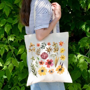 May include: A white canvas tote bag with a floral design. The flowers are in shades of yellow, orange, red, and pink. The bag has two long handles.