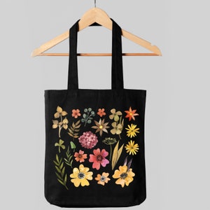 May include: Black tote bag with a floral design. The bag features a variety of flowers in shades of yellow, orange, pink, and red, as well as green leaves and stems. The flowers are arranged in a random pattern on the bag.
