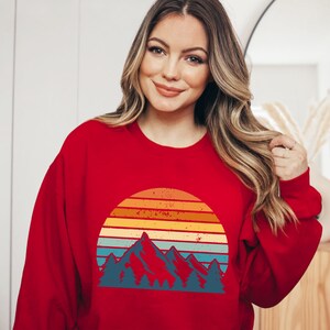 May include: Red sweatshirt featuring a vintage-style graphic of mountains and a sunset. The graphic is in shades of orange, yellow, blue, and green.