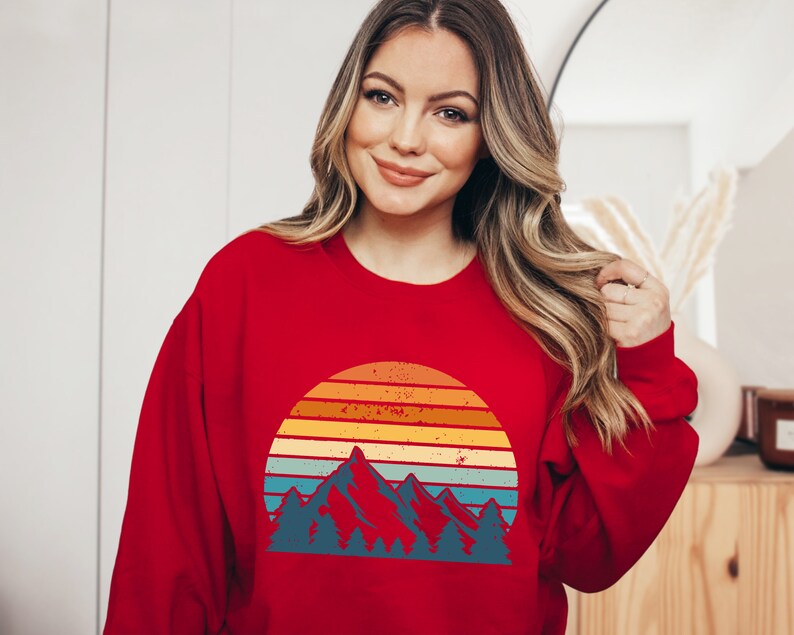 May include: Red sweatshirt featuring a vintage-style graphic of mountains and a sunset. The graphic is in shades of orange, yellow, blue, and green.