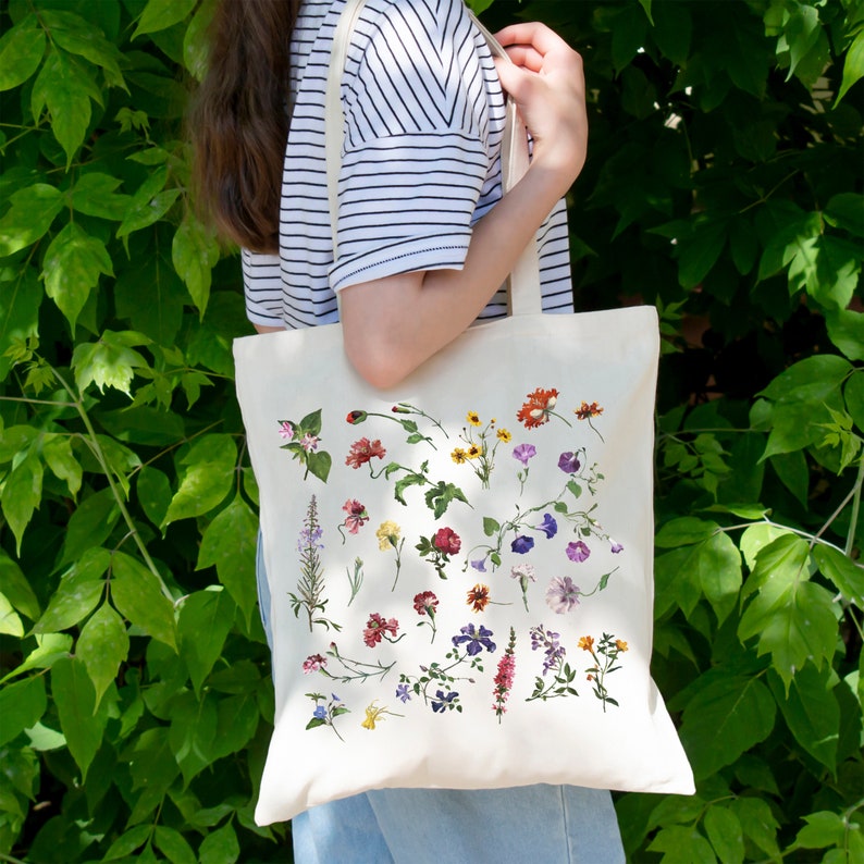 Peut inclure: Un sac fourre-tout en toile blanche avec un imprim&eacute; floral repr&eacute;sentant diff&eacute;rentes fleurs dans des tons de rose, violet, jaune et rouge. Les fleurs sont dispos&eacute;es de mani&egrave;re al&eacute;atoire sur le sac.