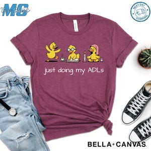 May include: A maroon t-shirt with a graphic of three yellow ducklings engaged in different activities. The text "just doing my ADLs" is printed below the ducks. The shirt is made of a soft material and is from the brand Bella+Canvas.