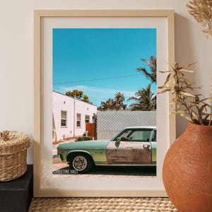 May include: A framed photograph of a vintage green car parked in front of a white building with a blue sky and palm trees in the background. The photo is signed "Christine Hall" in the bottom left corner.
