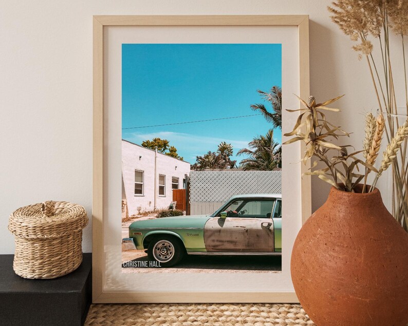 May include: A framed photograph of a vintage green car parked in front of a white building with a blue sky and palm trees in the background. The photo is signed "Christine Hall" in the bottom left corner.
