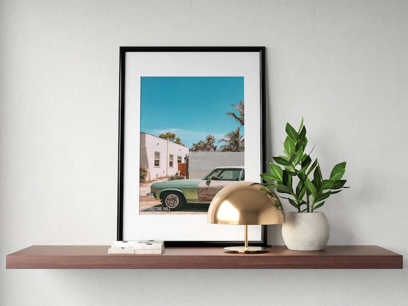 May include: A framed photograph of a vintage green car parked in front of a white building with a blue sky and palm trees in the background. A gold lamp with a dome-shaped shade sits on a wooden shelf with a potted plant.