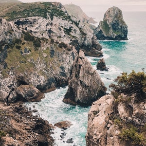 May include: A rocky coastline with a dramatic cliff face and crashing waves. The water is a vibrant blue and the sky is overcast. The cliffs are a mix of gray and brown.