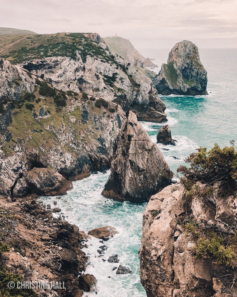 May include: A rocky coastline with a dramatic cliff face and crashing waves. The water is a vibrant blue and the sky is overcast. The cliffs are a mix of gray and brown.