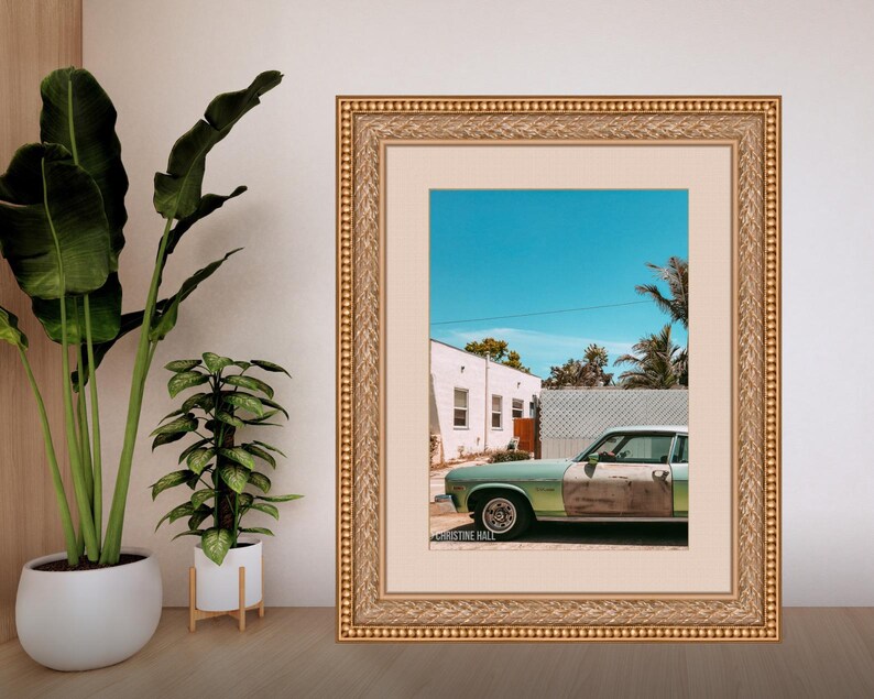 May include: A framed photograph of a vintage green car parked in front of a white house with a blue sky and palm trees in the background. The photo is signed "Christine Hall".