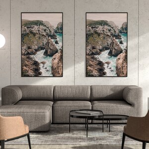 May include: Two framed landscape photographs of a rocky coastline with turquoise water. The photos are hanging on a light gray wall above a gray sectional sofa with two matching armchairs. The room is decorated with a white lamp and a vase.