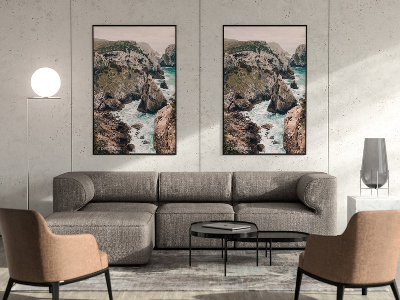 May include: Two framed landscape photographs of a rocky coastline with turquoise water. The photos are hanging on a light gray wall above a gray sectional sofa with two matching armchairs. The room is decorated with a white lamp and a vase.