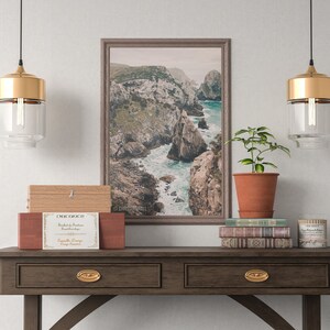 May include: A wooden console table with two drawers and gold hardware. The table is decorated with a framed photograph of a rocky coastline, a potted plant, a stack of books, and a candle. The text on the candle reads "The Little Branch Candle Co."