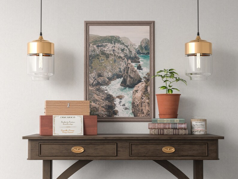 May include: A wooden console table with two drawers and gold hardware. The table is decorated with a framed photograph of a rocky coastline, a potted plant, a stack of books, and a candle. The text on the candle reads "The Little Branch Candle Co."