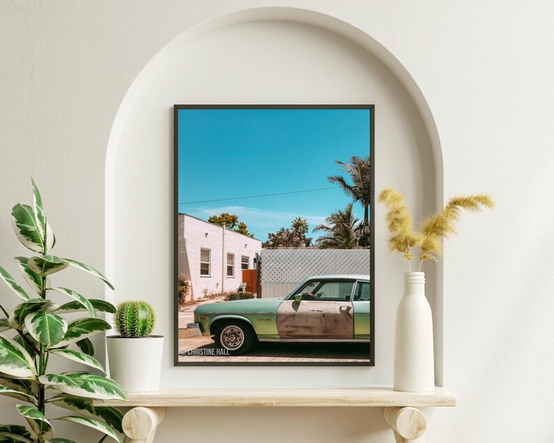 May include: A vintage green car parked in front of a white building with a blue sky and palm trees in the background. The car has rust on the side and the text "CHRISTINE HALL" is visible on the bottom of the image.