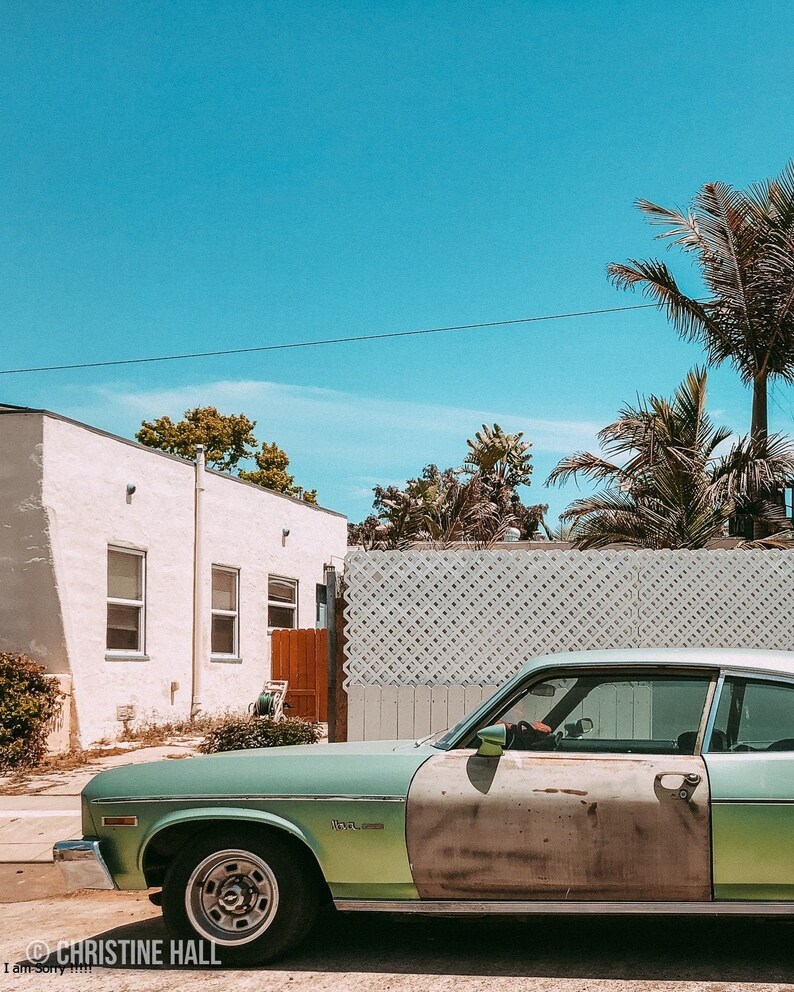 May include: A vintage green car parked in front of a white building with a wooden fence. The car has a faded paint job and a dent on the driver's side door. The car is parked on a street with a blue sky in the background.