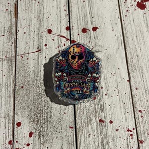 May include: A decorative badge featuring a colorful illustration of a hockey mask with the text "WELCOME TO CAMP CRYSTAL LAKE". The badge has a glittery border and is set against a distressed wood background with red splatters.