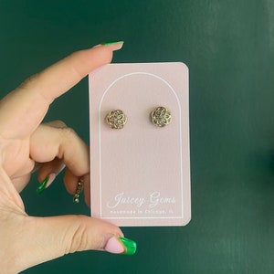 May include: A pair of gold-toned earrings with a delicate floral design. The earrings are displayed on a pink card with the text "Juicy Gems Handmade in Chicago, IL".