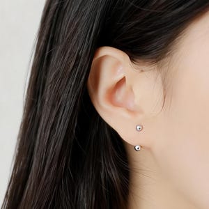 May include: Close-up of a person's ear with two small, silver ball earrings. The earrings are simple and minimalist, with one ball visible in front of the ear and another behind the earlobe. The person has long, dark hair.