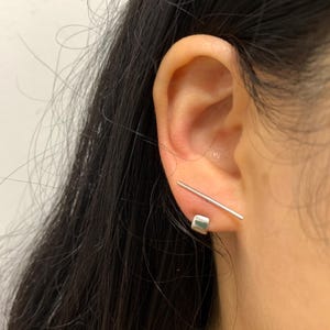 May include: A close-up of silver earrings. One earring has a small square stud and a straight bar across the ear. The other earring is a straight bar. The earrings are worn on a person's ear.