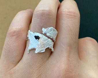 May include: A silver ring with a textured, organic design featuring a black stone. The ring is worn on a person's finger.