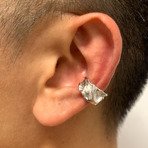 May include: A silver ear cuff with a textured, organic design. The cuff is worn on the ear and has a flowing, sculptural shape. The jewellery piece is made of a shiny, metallic material.
