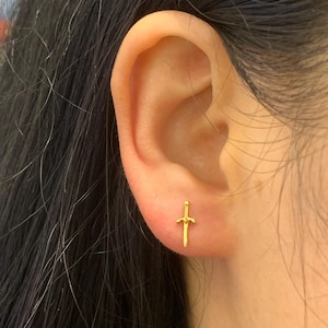 May include: A close-up of an ear with a small, gold-coloured sword-shaped stud earring. The earring is a detailed miniature of a sword, with a hilt and blade. The earring is set against the skin of the ear.