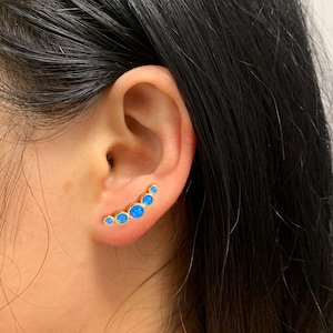 Puede incluir: Un pendiente trepador dorado con cinco piedras de ópalo azul. Las piedras están dispuestas en una línea curva, creando un aspecto elegante y moderno. El pendiente está diseñado para usarse a lo largo del lóbulo de la oreja.
