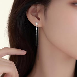 May include: A close-up of a woman's ear adorned with delicate silver earrings. Each earring features a small butterfly stud and two long, dangling chains. The earrings are a simple yet elegant accessory.