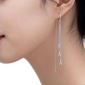 Könnte beinhalten: Silberne Ohrhänger mit drei tropfenförmigen Anhängern. Die Ohrringe sind lang und hängen herab, mit einem einfachen Hakenverschluss. Das Design ist elegant und minimalistisch, geeignet für den Alltag oder besondere Anlässe.