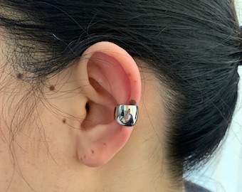 Könnte beinhalten: Ein glänzender, silberner Ohrschmuck wird am oberen Ohr getragen. Der Schmuck hat eine abgerundete, zylindrische Form und reflektiert die Umgebung. Das Ohr und die Haare der Person sind sichtbar.