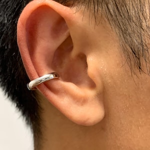 Puede incluir: Un ear cuff plateado se lleva en la parte superior de la oreja. El ear cuff tiene un diseño sencillo y redondeado, y parece estar hecho de metal pulido. El ear cuff es una pieza de joyería moderna.