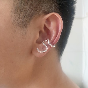 May include: A close-up of a person's ear with three silver ear cuffs. The cuffs are made of thin, twisted metal and are designed to hug the ear.