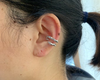 Puede incluir: Un pendiente de aro de plata con un diseño retorcido. El aro se lleva en la parte superior de la oreja.