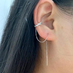 Może przedstawiać: Srebrny nausznik z zygzakowatym wzorem i wiszącym kolczykiem z łańcuszkiem. Nausznik jest ozdobiony małymi, błyszczącymi kamieniami. Kolczyk ma delikatny łańcuszek, który zwisa z ucha.